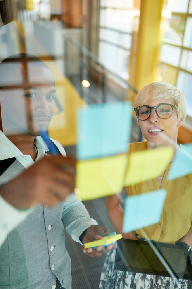 Two creative millenial small business owners working on social media strategy brainstorming using adhesive notes in windows-1